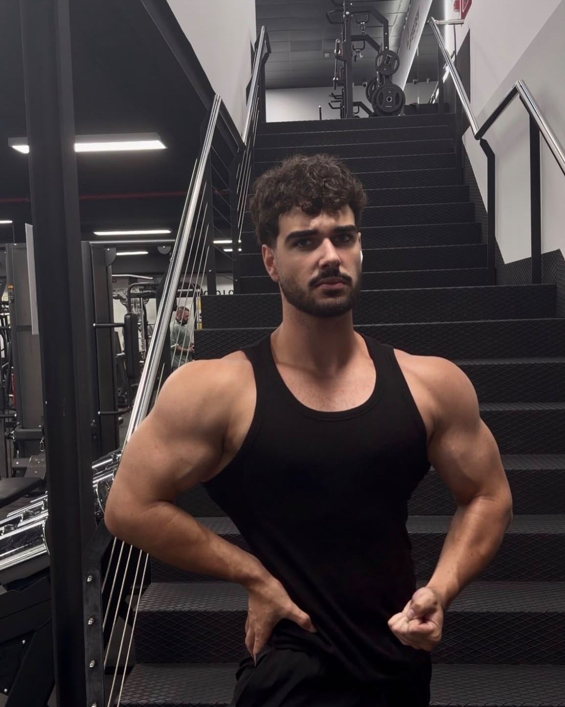 Aaron, coach de transformación física, posando en camiseta de tirantes negra en las escaleras de un gimnasio